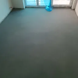 Świeżo wylana, szara posadzka jastrychowa w pomieszczeniu z oknem zabezpieczonym folią budowlaną.