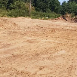 Przygotowany teren pod składowanie, widoczne ślady opon po maszynach budowlanych, w tle zieleń drzew.