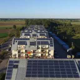 Widok z lotu ptaka na osiedle z panelami słonecznymi na dachach, otoczone zielenią i polami w oddali, w słoneczny dzień.