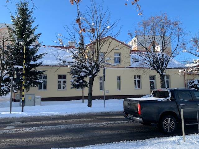 Dwupiętrowy budynek w kolorze kremowym z pomarańczowym dachem pokrytym śniegiem, widoczny z ulicy w zimowej scenerii, zaparkowany czarny pickup.