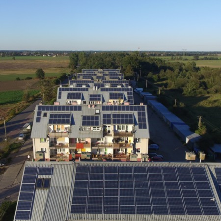 Widok z lotu ptaka na osiedle z panelami słonecznymi na dachach, otoczone zielenią i polami w oddali, w słoneczny dzień.