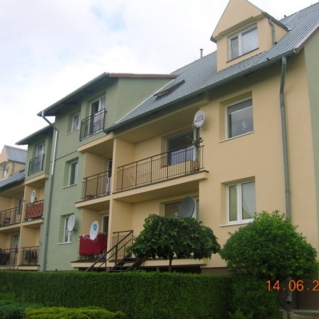 Fasada budynku wielorodzinnego z balkonami, pomalowana na żółto i jasnozielono, z antenami satelitarnymi na ścianach i zadbanym żywopłotem przed budynkiem.