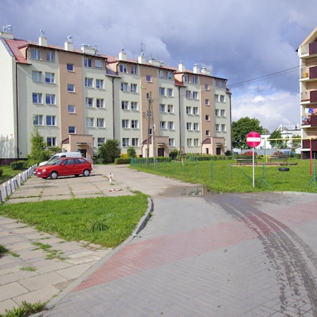 Widok na wielorodzinny budynek mieszkalny z placem zabaw, czerwonym samochodem i znakiem zakazu wjazdu.