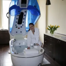 Uśmiechnięta kobieta w białym szlafroku siedzi przy otwartej kapsule SPA z hydromasażem, w tle bukiet żółtych tulipanów i czarne lampy.