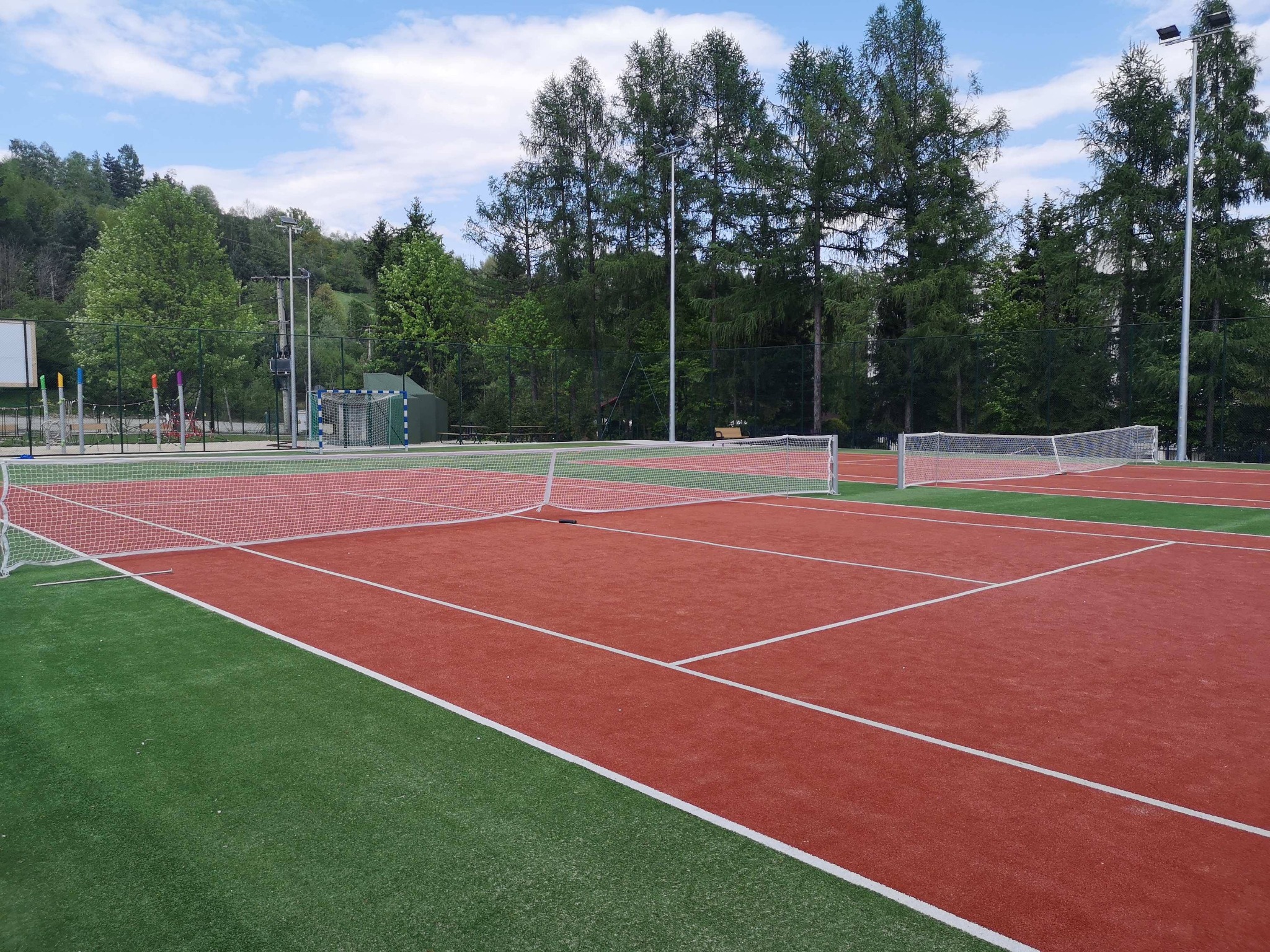 Czerwono-zielony kort tenisowy z siatkami na zewnątrz, otoczony drzewami i wzgórzami w tle.