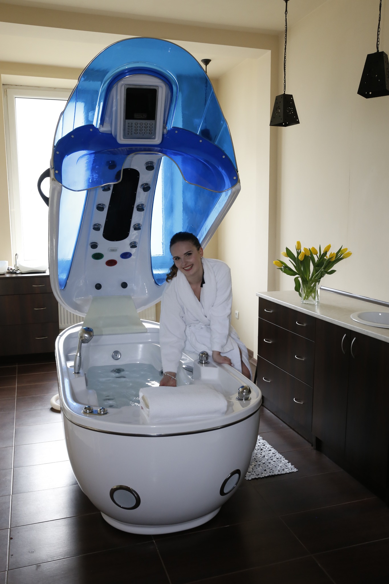 Uśmiechnięta kobieta w białym szlafroku siedzi przy otwartej kapsule SPA z hydromasażem, w tle bukiet żółtych tulipanów i czarne lampy.