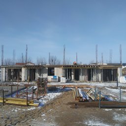 Ingbau Spólka z Ograniczoną Odpowiedzialnością Sp. z o.o - Surowy stan budowy domu z betonu komórkowego w zimowej scenerii, widoczne szalunki, zbrojenia i materiały budowlane, w tle pracownik w pomarańczowej kamizelce na dachu.