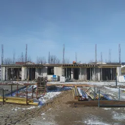 Surowy stan budowy domu z betonu komórkowego w zimowej scenerii, widoczne szalunki, zbrojenia i materiały budowlane, w tle pracownik w pomarańczowej kamizelce na dachu.