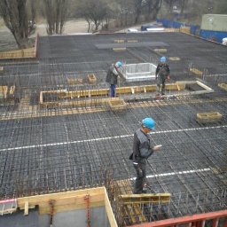 Ingbau Spólka z Ograniczoną Odpowiedzialnością Sp. z o.o - Prace zbrojarskie na budowie, widoczne siatki zbrojeniowe, szalunki i pracownicy w kaskach na tle miejskiego krajobrazu.