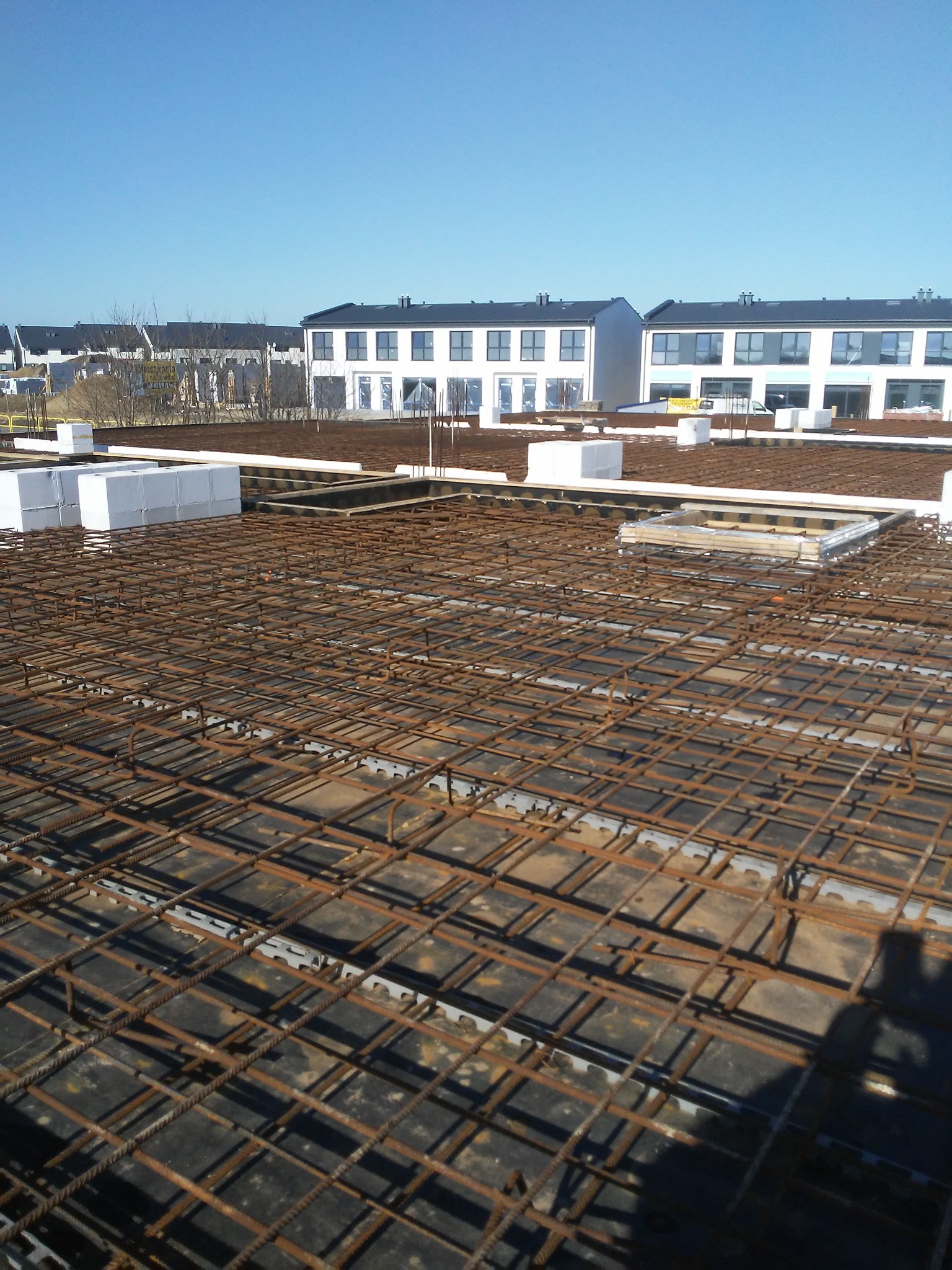 Zbrojenie stropu budynku wielorodzinnego z widoczną siatką stalową, elementami szalunków i izolacji termicznej, w tle budynki w stanie surowym zamkniętym.