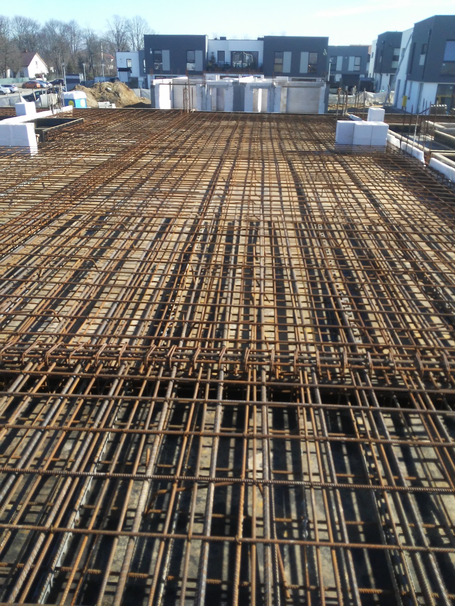 Zbrojenie płyty fundamentowej domu jednorodzinnego, widoczne siatki z prętów stalowych i białe bloczki betonowe w tle.