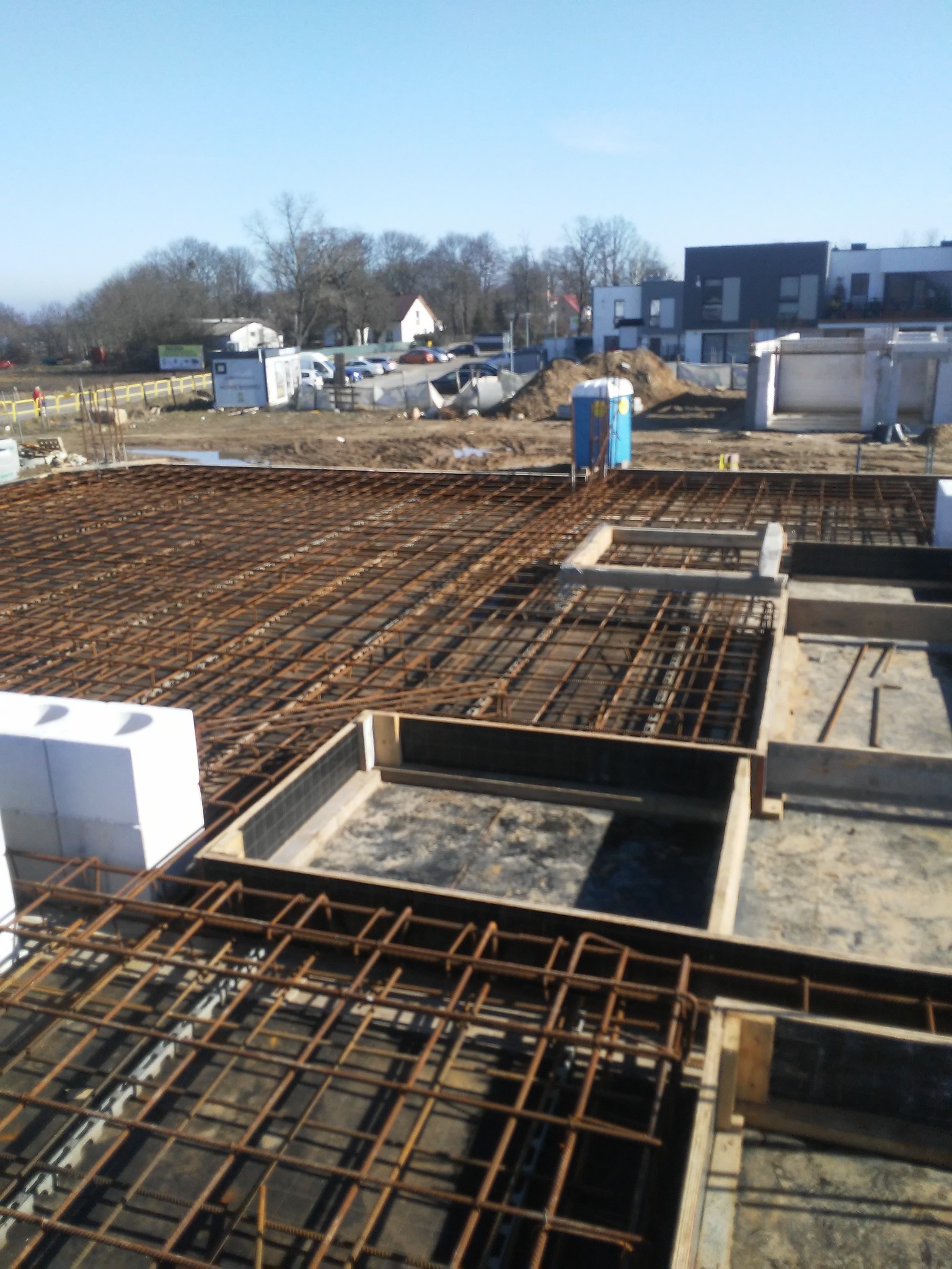 Zbrojenie fundamentów domu jednorodzinnego z widocznymi szalunkami i stosem białych bloczków styropianowych, na tle placu budowy i zabudowy mieszkalnej.