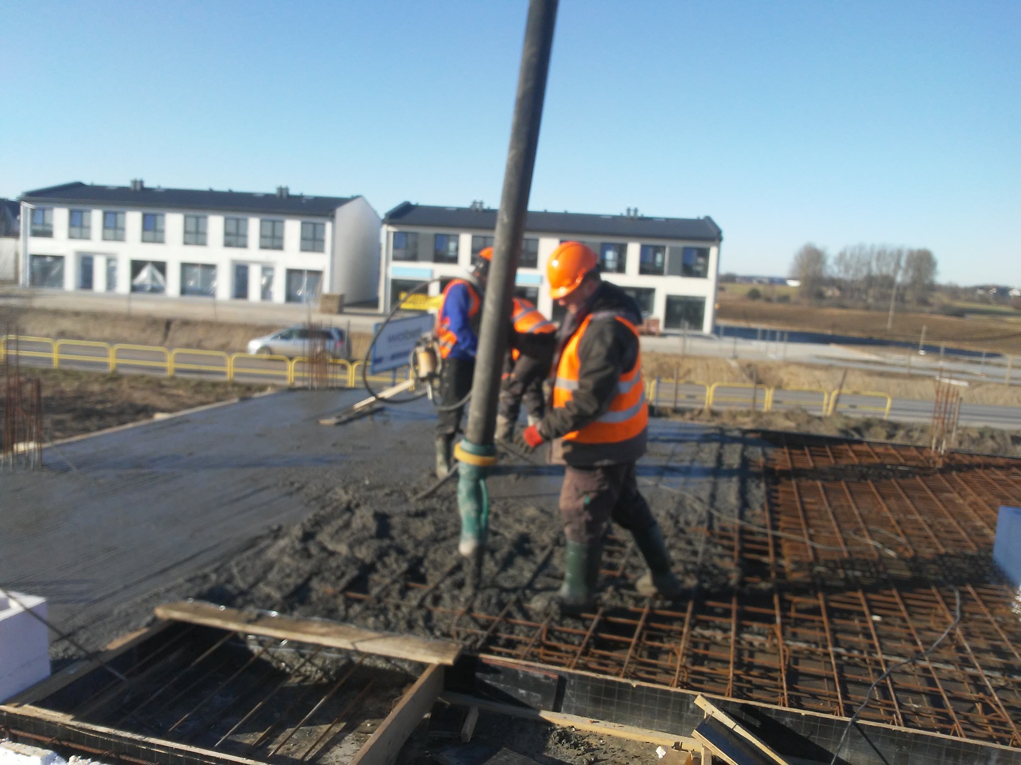 Wylewanie betonu na zbrojenie fundamentów domu jednorodzinnego, widoczni pracownicy w odblaskowych kamizelkach i kaskach, w tle budynki w stanie surowym otwarte.