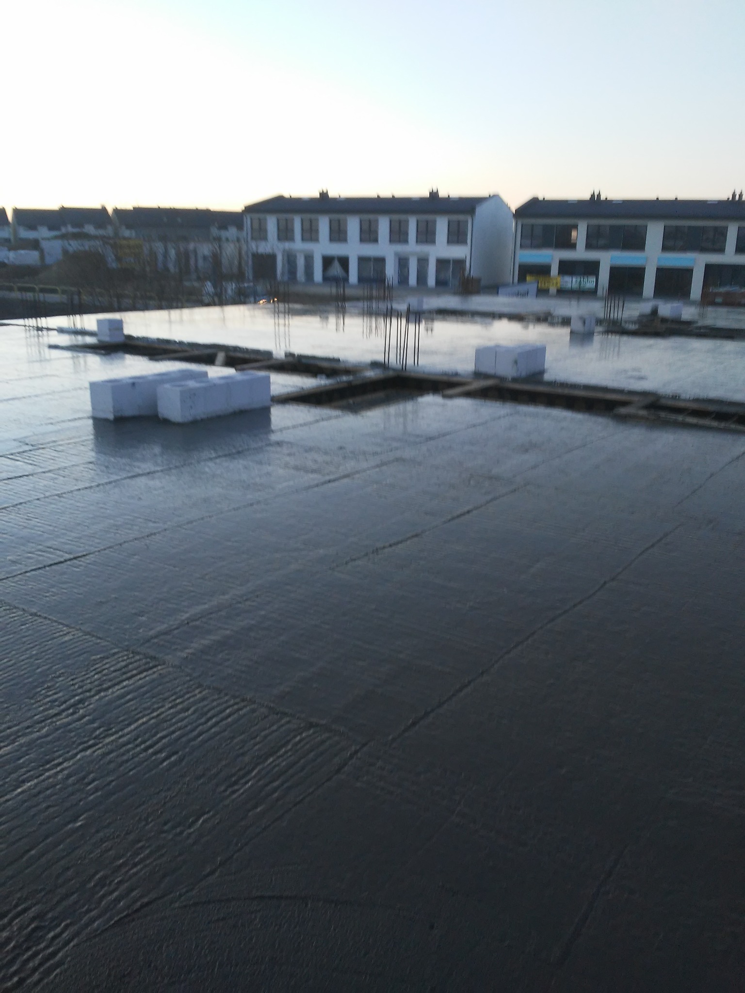 Świeżo wylany beton na stropie budynku w trakcie budowy, widoczne bloczki betonowe i wystające pręty zbrojeniowe, w tle budynki mieszkalne.