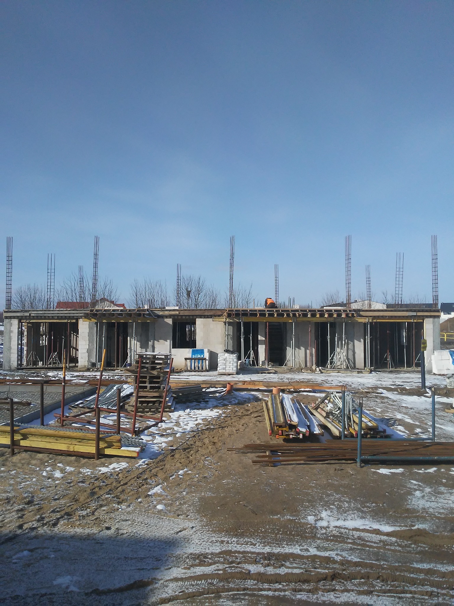 Surowy stan budowy domu z betonu komórkowego w zimowej scenerii, widoczne szalunki, zbrojenia i materiały budowlane, w tle pracownik w pomarańczowej kamizelce na dachu.