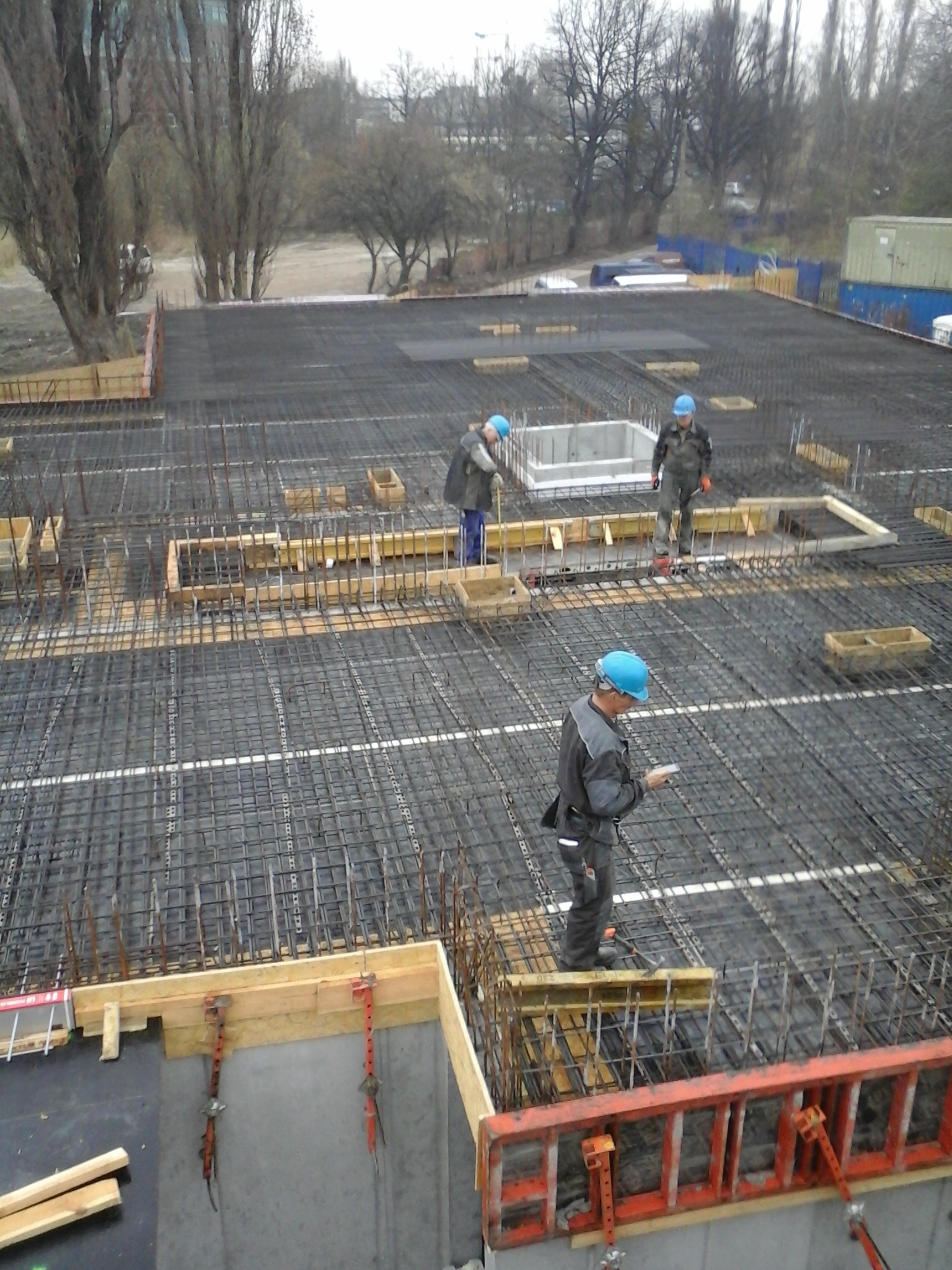 Prace zbrojarskie na budowie, widoczne siatki zbrojeniowe, szalunki i pracownicy w kaskach na tle miejskiego krajobrazu.