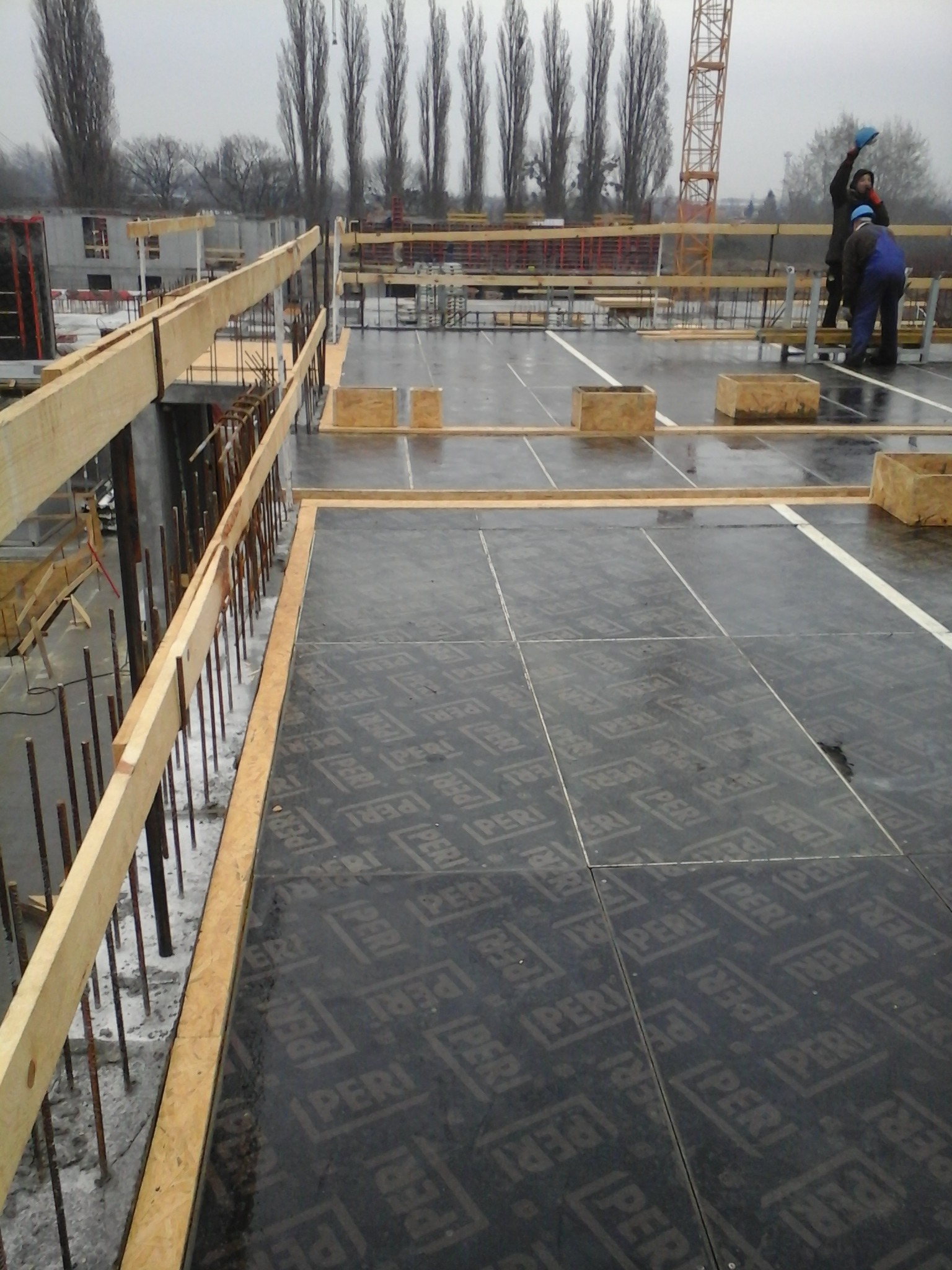 Budowa stropu z drewnianymi belkami i deskami szalunkowymi z nadrukiem PERI, widoczni robotnicy w oddali oraz żuraw budowlany.