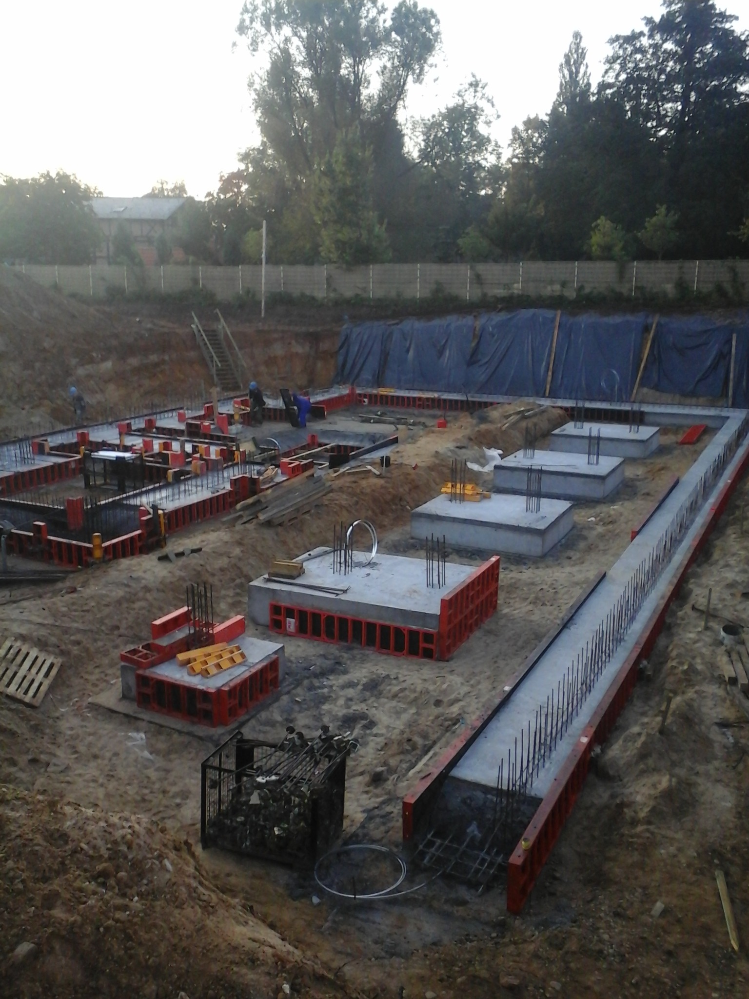 Wykop pod fundamenty domu z widocznymi zbrojeniami i szalunkami w kolorze czerwonym, przygotowany do zalania betonem, w tle drzewa i budynek.