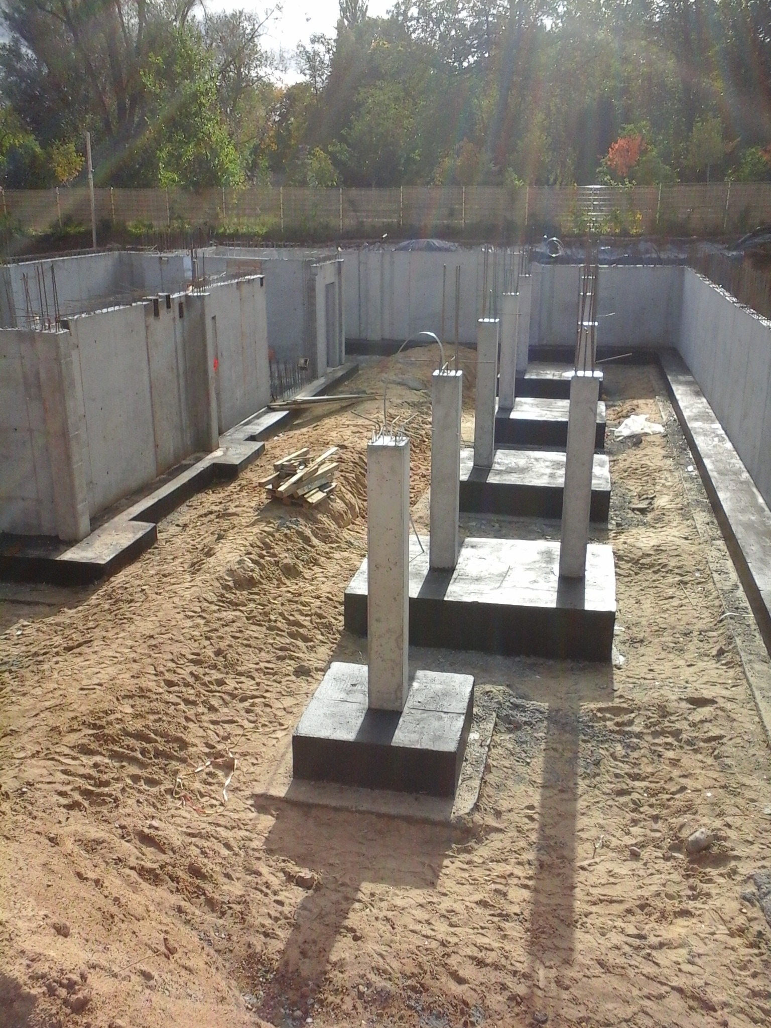 Fundamenty domu w trakcie budowy z widocznymi ścianami piwnicznymi i betonowymi słupami, teren zasypany piaskiem.