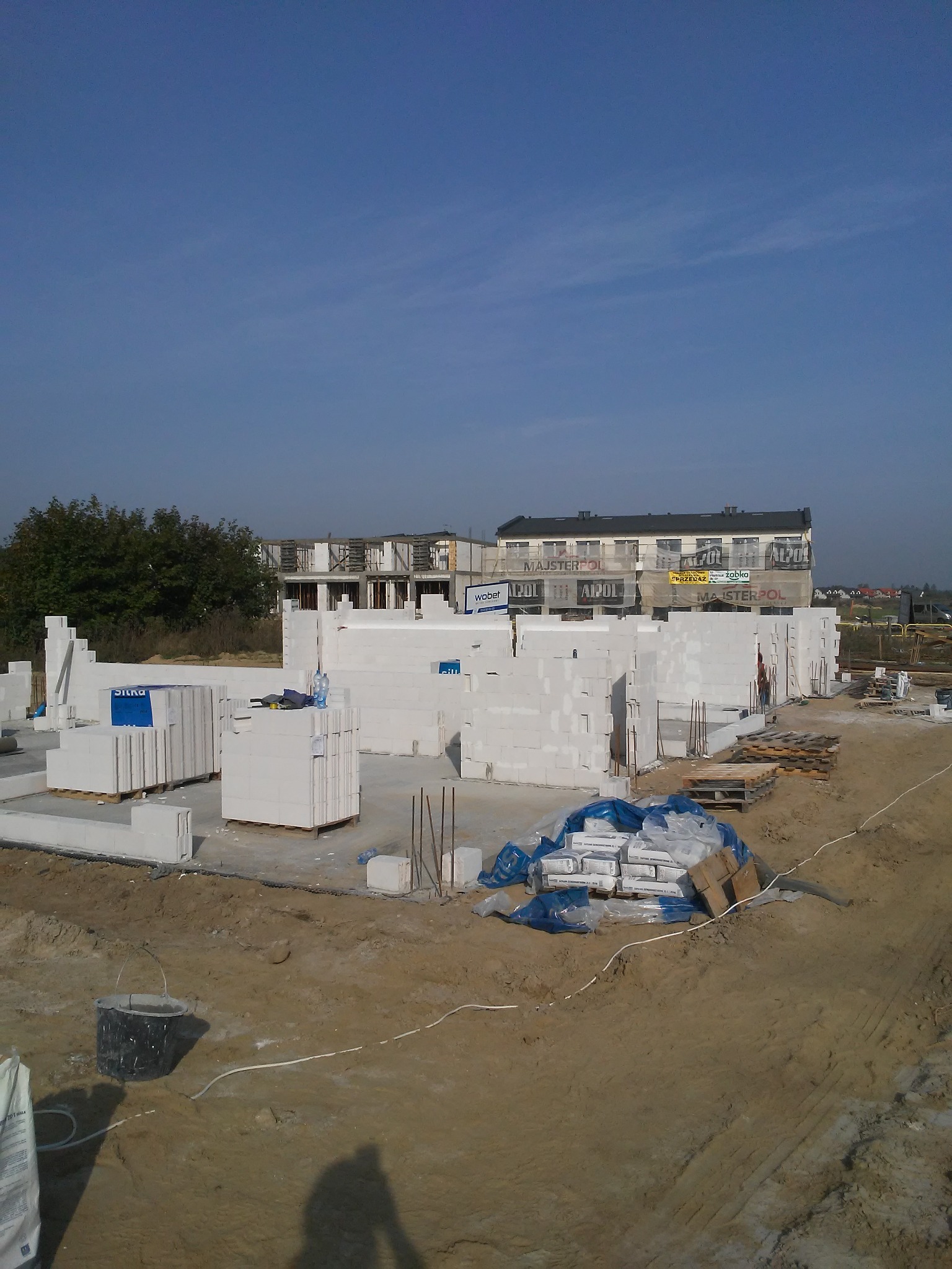 Wznoszenie ścian z białych bloczków na placu budowy domu wielorodzinnego, widoczne materiały budowlane i zarys sylwetki fotografa.