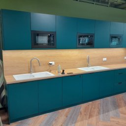 Netkam Anna Suszka - Turkusowa zabudowa kuchenna z drewnianym blatem i wbudowanymi mikrofalówkami, widok na dwie umywalki z bateriami i akcesoria na blacie, podłoga imitująca drewno ułożona w jodełkę.