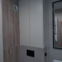 Netkam Anna Suszka - Biała szafka łazienkowa podwieszana z ukrytym systemem montażu, widoczny fragment umywalki i lustra z podświetleniem LED, na ścianie imitacja drewna i szara płytka.