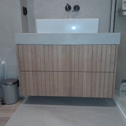 Netkam Anna Suszka - Łazienka z drewnianą szafką pod umywalką, szara armatura i płytki, widoczny kosz na śmieci i fragment toalety.