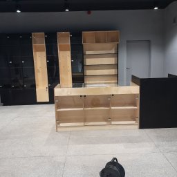 Netkam Anna Suszka - Wnętrze sklepu w trakcie montażu: widoczne regały z jasnego drewna z przeszklonymi frontami, czarne szafki z połyskiem w tle, narzędzia w czerwonym pojemniku i zwinięty przedłużacz na podłodze.