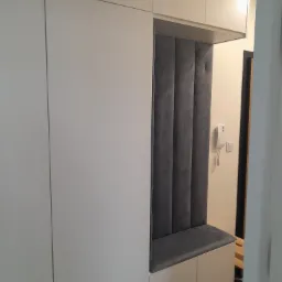 Biała szafa wnękowa z siedziskiem i tapicerowanym panelem ściennym w kolorze szarym, w tle widoczny domofon i fragment schodów.