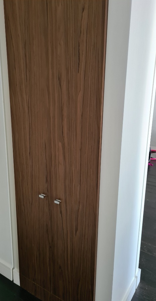 Wysoka szafa wnękowa z drewnianymi drzwiami o widocznym usłojeniu, wkomponowana w białą ścianę.