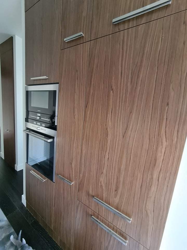 Zabudowa kuchenna z forniru orzechowego z wbudowanym piekarnikiem i mikrofalówką, widoczne podłużne metalowe uchwyty.