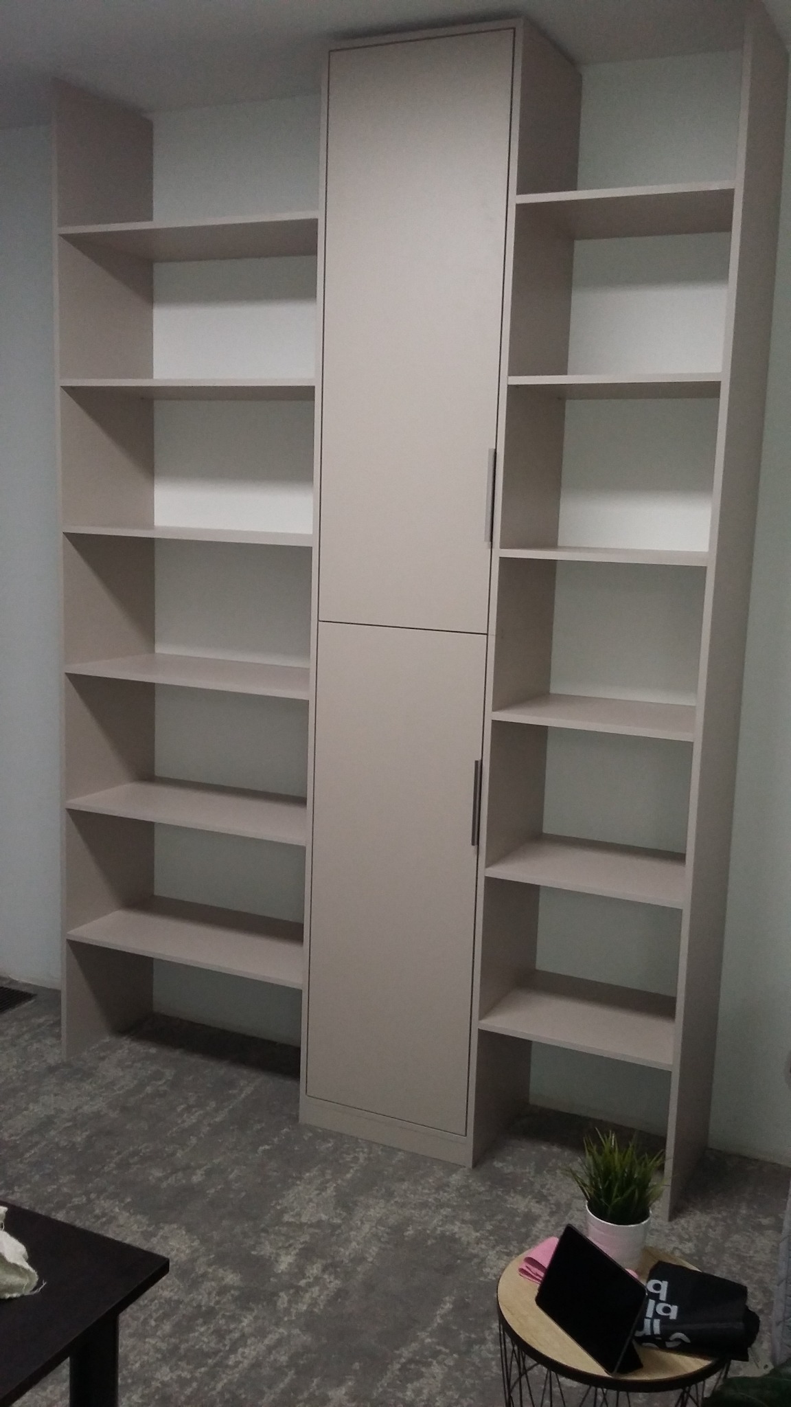 Beżowa szafa wnękowa z otwartymi półkami po bokach, zlokalizowana w pomieszczeniu z szarą wykładziną dywanową, widoczny fragment stolika kawowego i stolika pomocniczego z laptopem i rośliną...