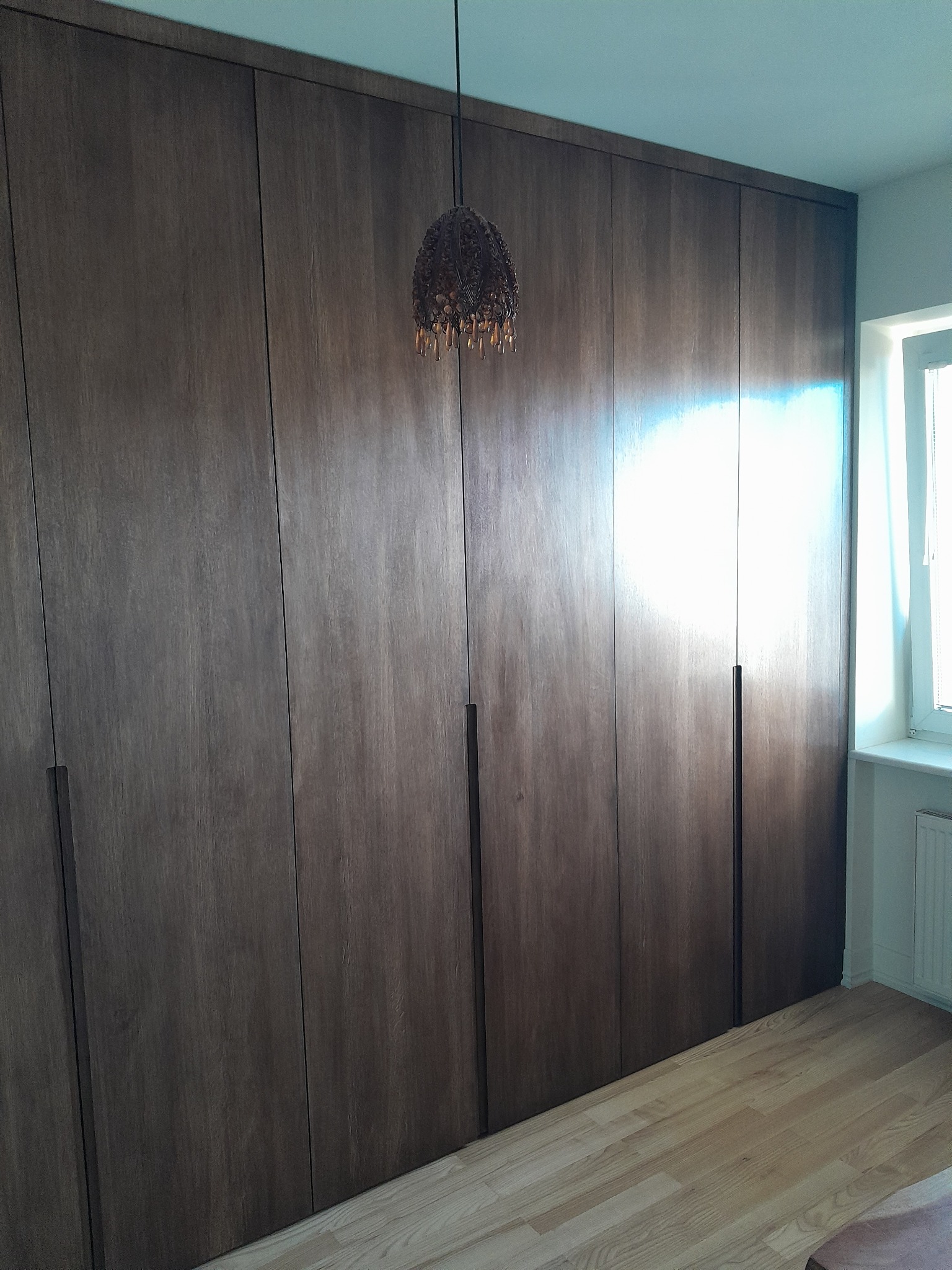 Duża, drewniana zabudowa garderoby od podłogi do sufitu, z dyskretnymi, pionowymi uchwytami, oświetlona wiszącą lampą z frędzlami. Parkiet na podłodze.