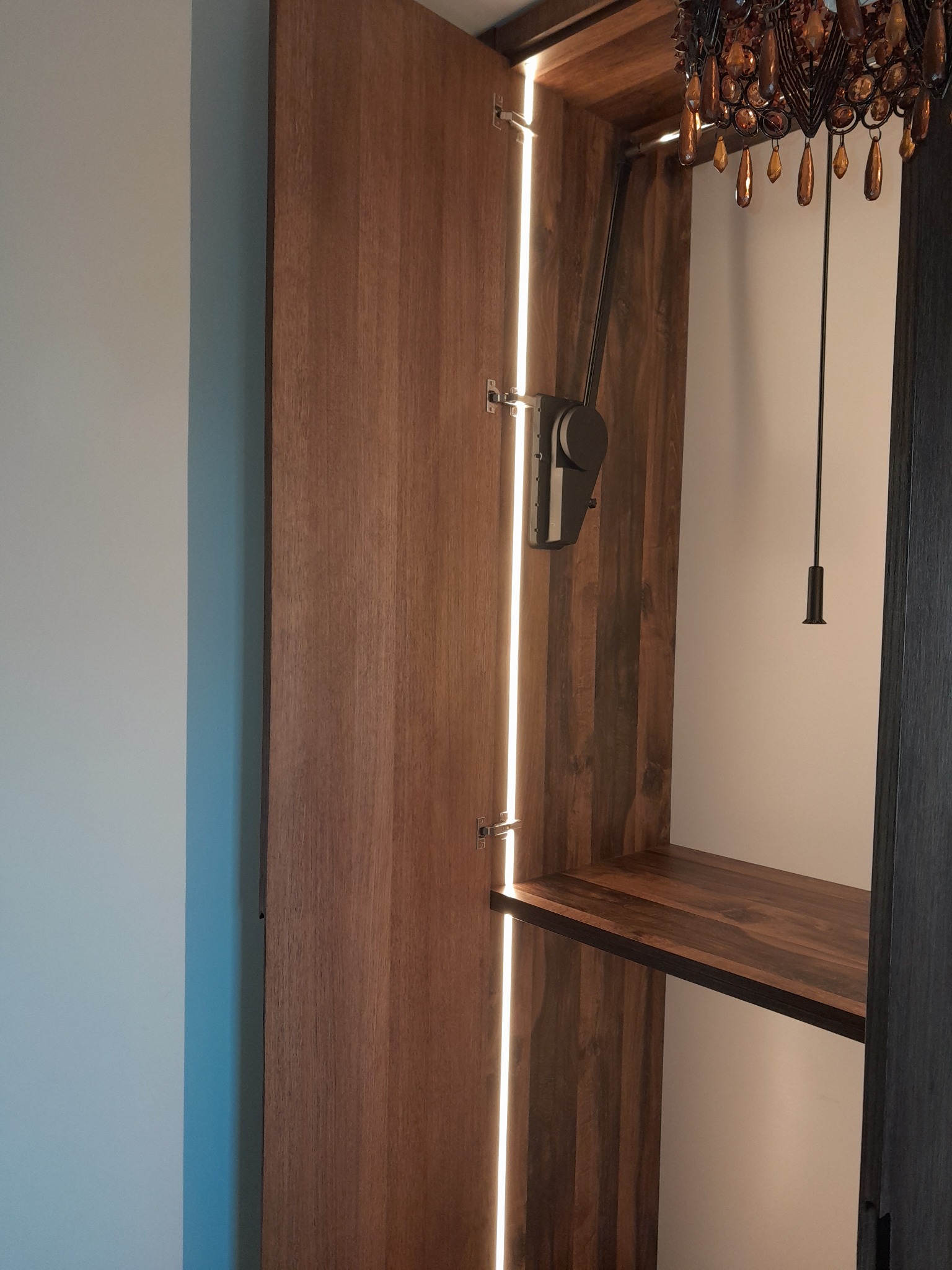 Otwarta drewniana szafa z wewnętrznym oświetleniem LED i półką, widoczny mechanizm opuszczania drążka na ubrania oraz fragment żyrandola.