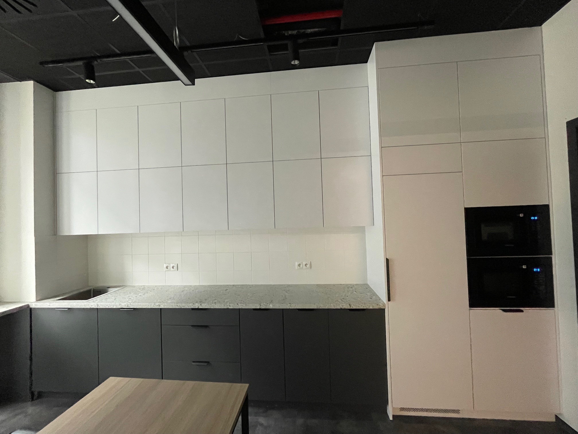 Nowoczesna kuchnia z szarymi szafkami dolnymi, jasnym blatem i białymi szafkami górnymi, zabudowana z piekarnikiem i mikrofalówką, oświetlenie punktowe na czarnym suficie.