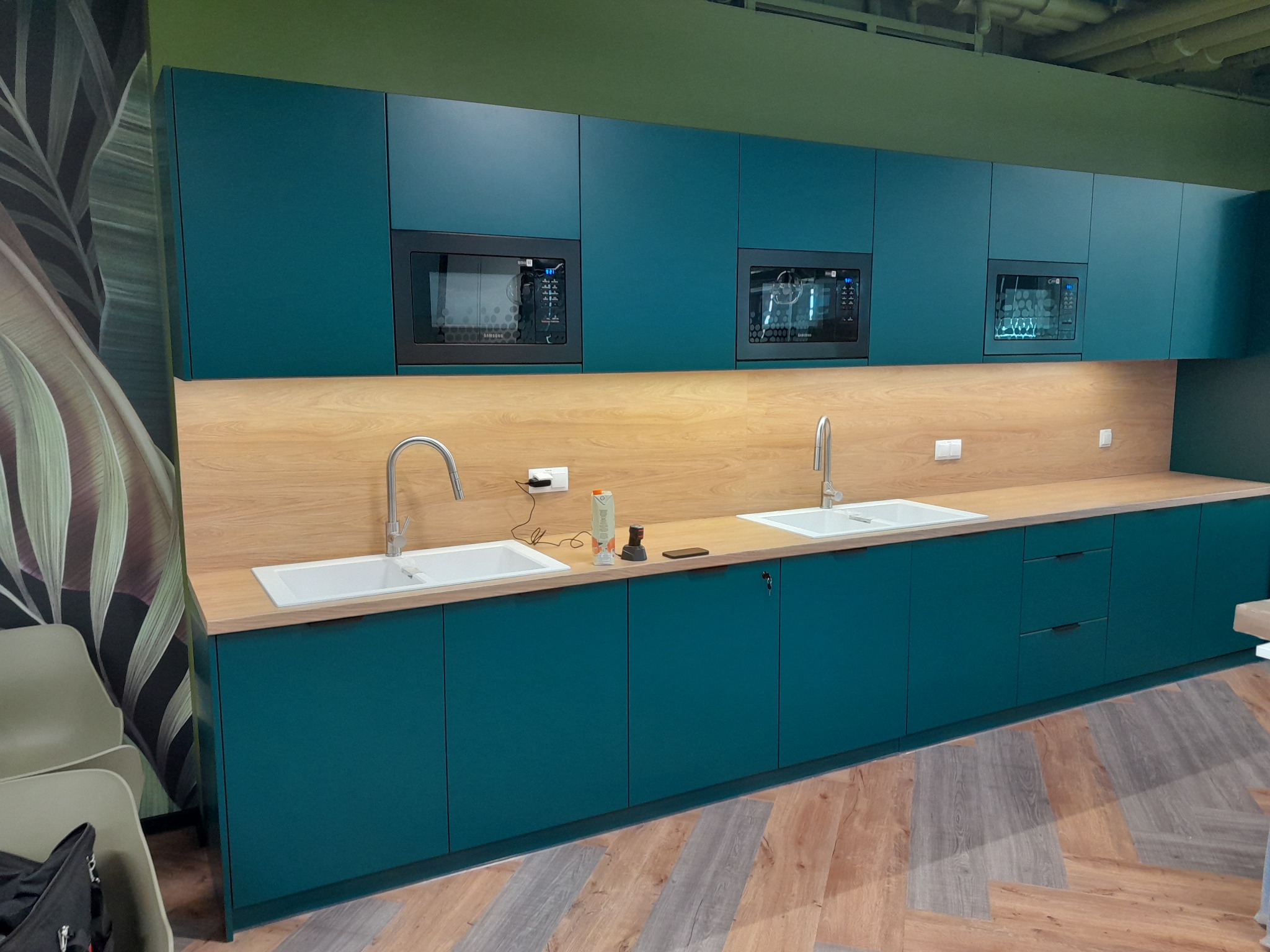Turkusowa zabudowa kuchenna z drewnianym blatem i wbudowanymi mikrofalówkami, widok na dwie umywalki z bateriami i akcesoria na blacie, podłoga imitująca drewno ułożona w jodełkę.