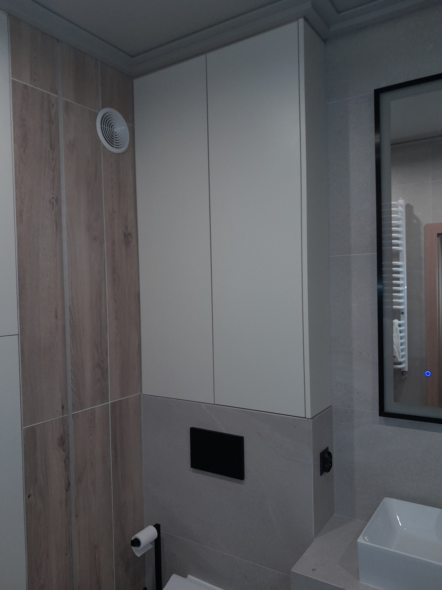 Biała szafka łazienkowa podwieszana z ukrytym systemem montażu, widoczny fragment umywalki i lustra z podświetleniem LED, na ścianie imitacja drewna i szara płytka.