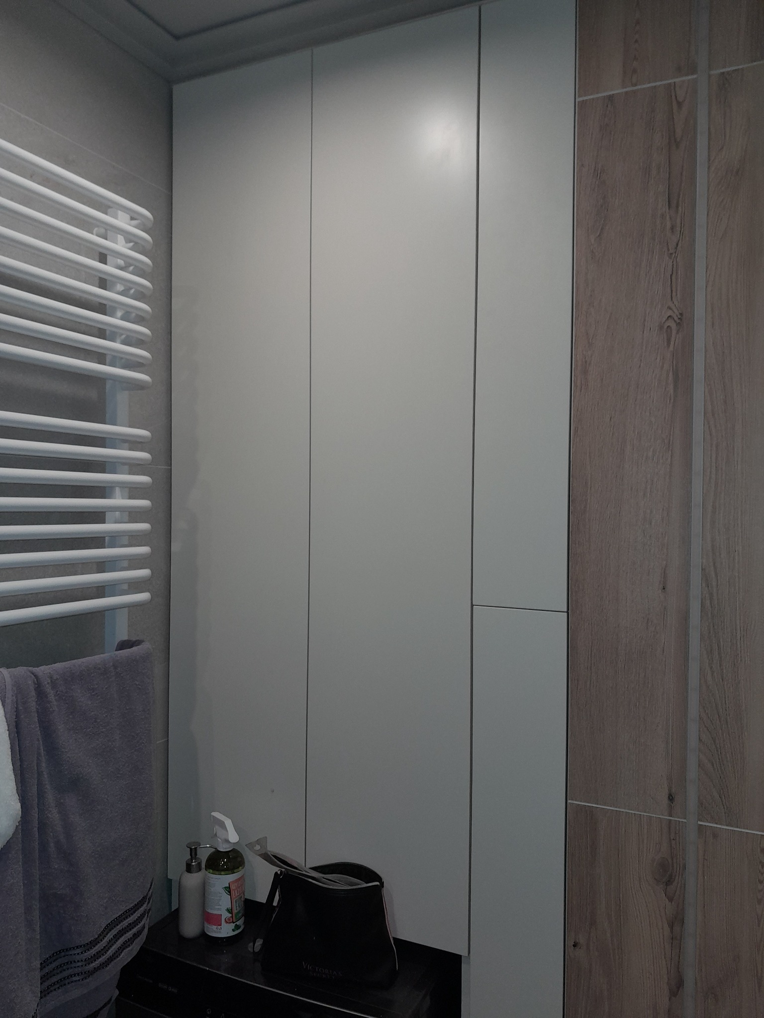 Biała, zabudowana szafa z drewnianymi akcentami obok grzejnika drabinkowego z powieszonym ręcznikiem i kosmetykami na pralce