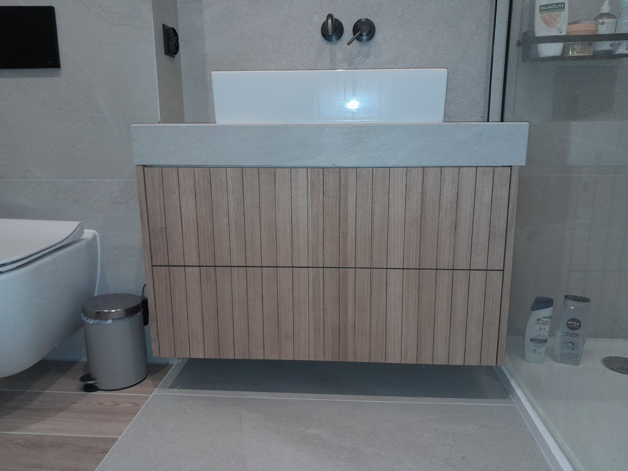 Łazienka z drewnianą szafką pod umywalką, szara armatura i płytki, widoczny kosz na śmieci i fragment toalety.