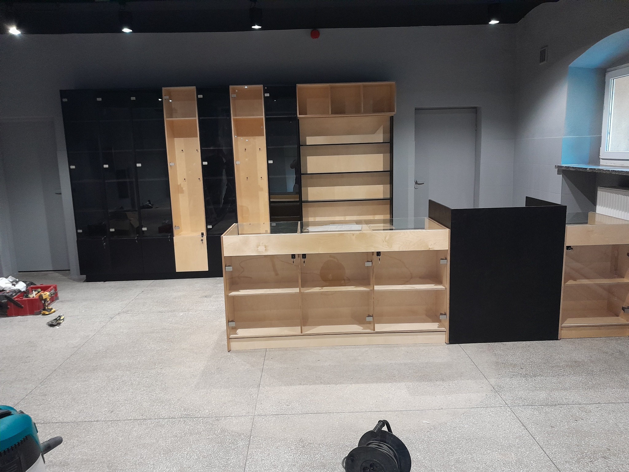 Wnętrze sklepu w trakcie montażu: widoczne regały z jasnego drewna z przeszklonymi frontami, czarne szafki z połyskiem w tle, narzędzia w czerwonym pojemniku i zwinięty przedłużacz na podłodze.