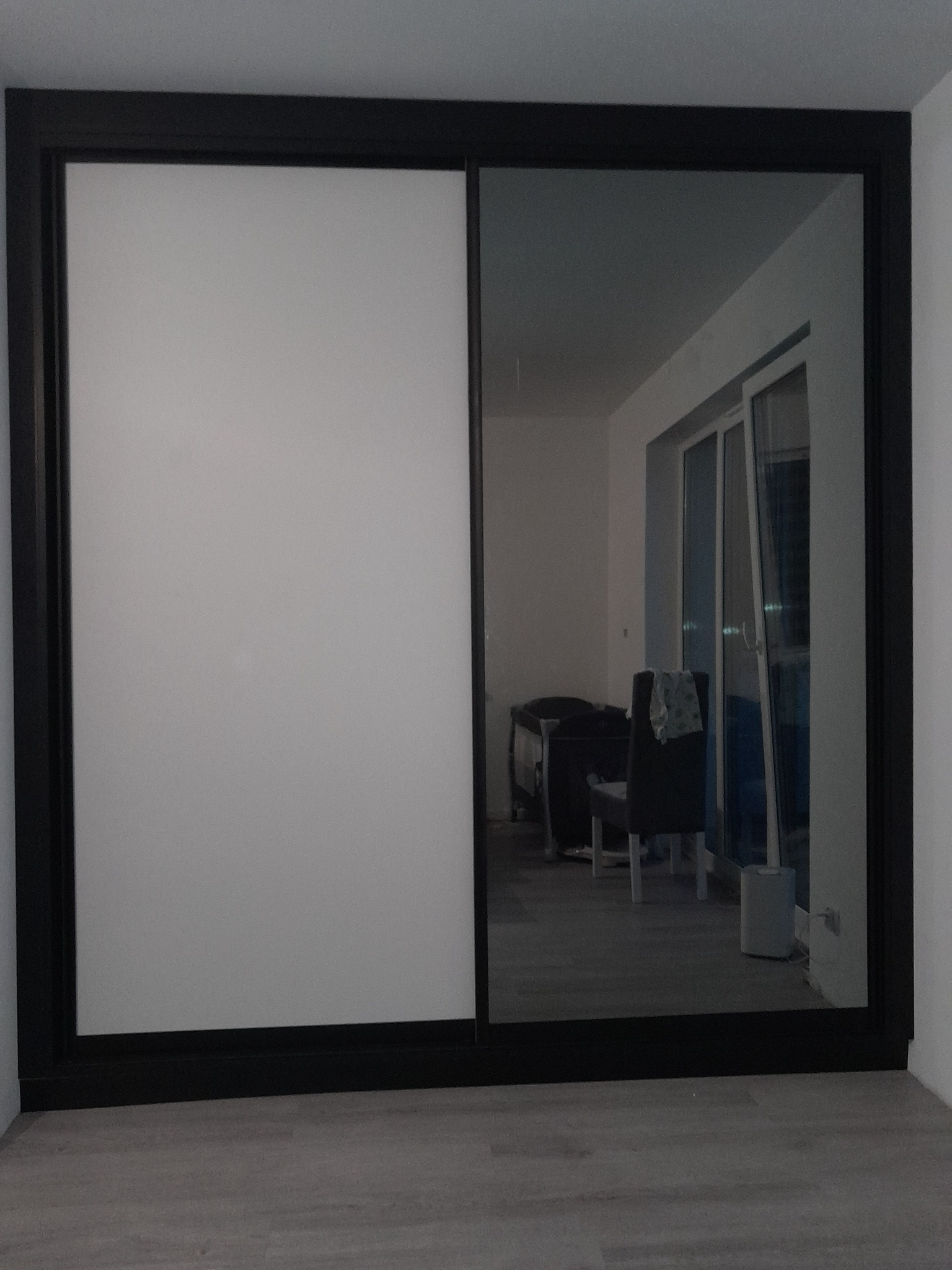 Czarna szafa wnękowa z przesuwnymi drzwiami, jedno skrzydło białe, drugie lustrzane, odbijające fragment pokoju z oknem balkonowym i meblami.