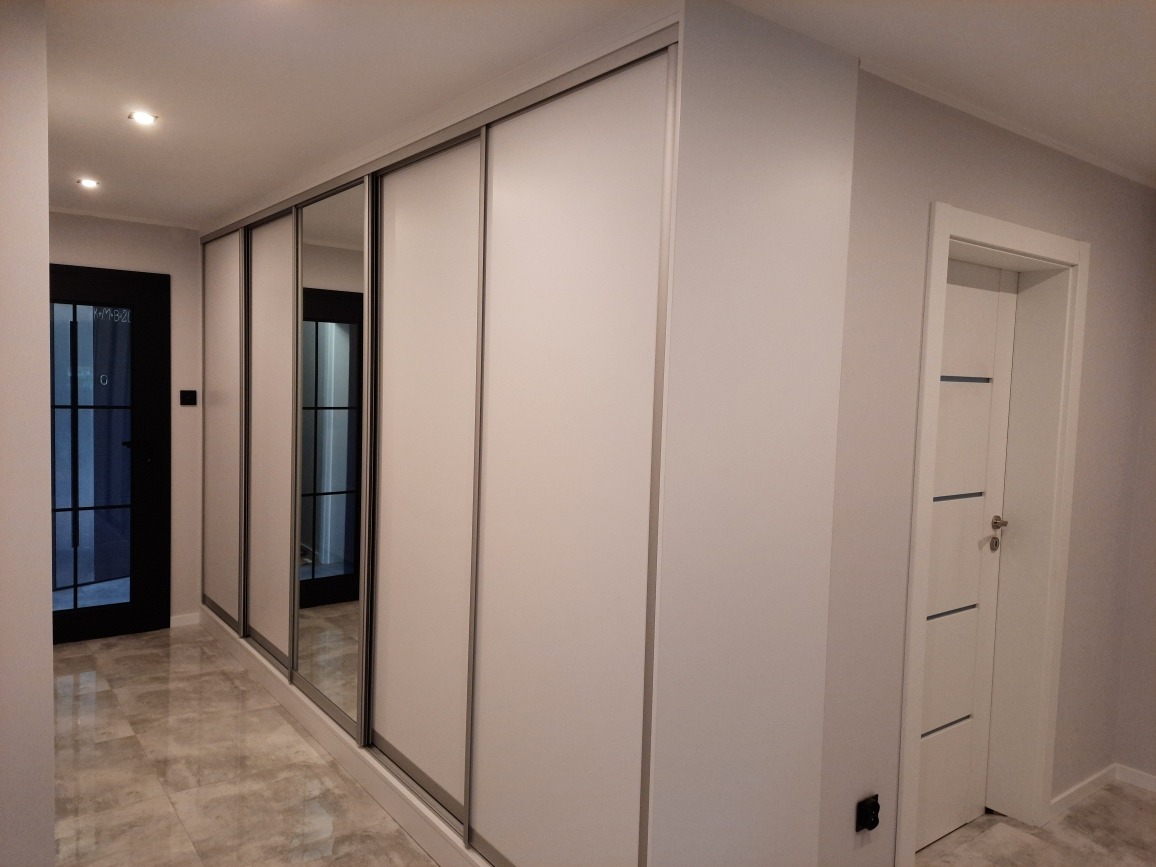 Biała szafa wnękowa z przesuwnymi drzwiami, w tym jedno lustrzane, w korytarzu z szarą podłogą i widocznymi drzwiami.