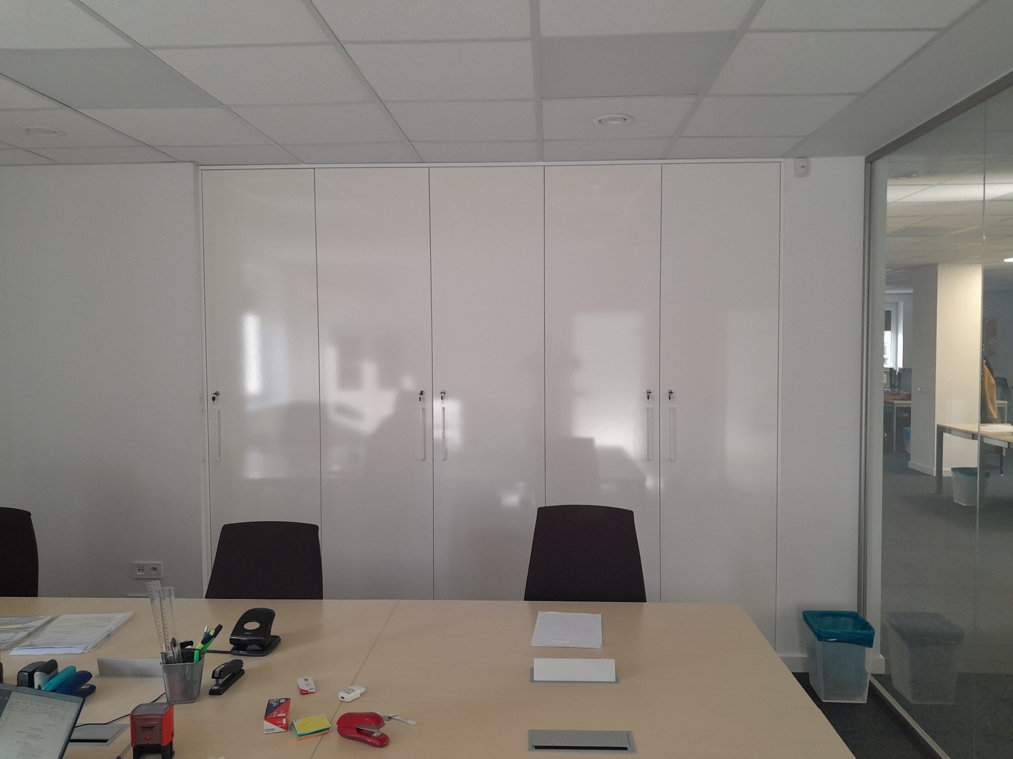 Biała szafa wnękowa z czterema drzwiami, zamykana na klucz, zamontowana w biurze, widoczna zza stołu konferencyjnego z krzesłami i biurowymi akcesoriami.