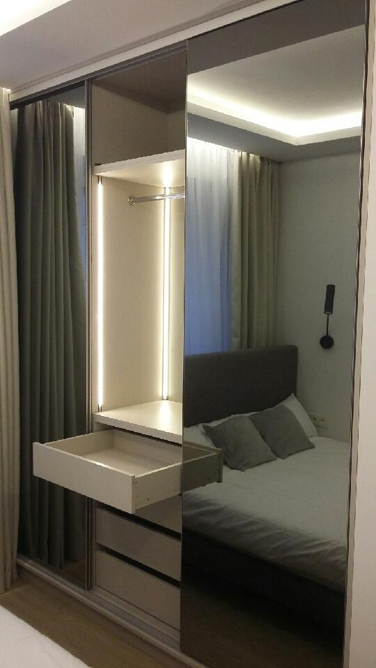 Wnętrze szafy z przesuwnymi drzwiami lustrzanymi, otwartą szufladą i oświetleniem LED, odbijające fragment sypialni z łóżkiem i szarą pościelą.