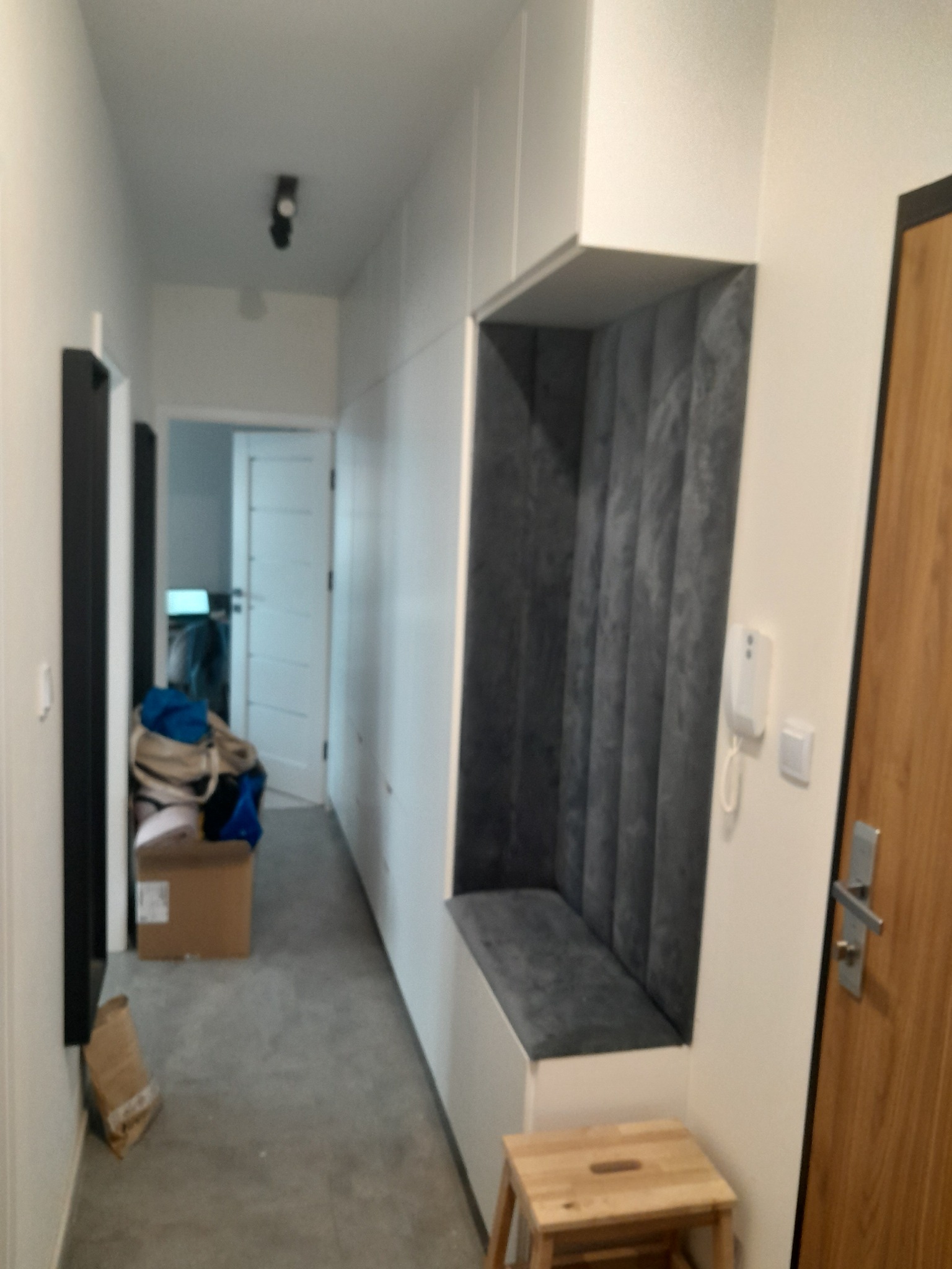 Długi korytarz z zabudowaną szafą wnękową z siedziskiem obitym szarą tapicerką, mały drewniany taboret i drzwi wejściowe.