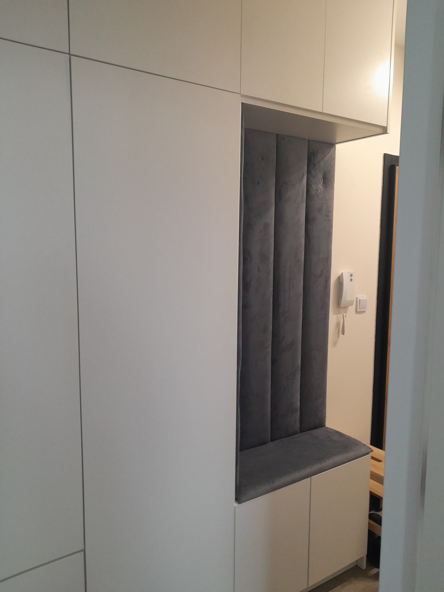 Biała szafa wnękowa z siedziskiem i tapicerowanym panelem ściennym w kolorze szarym, w tle widoczny domofon i fragment schodów.