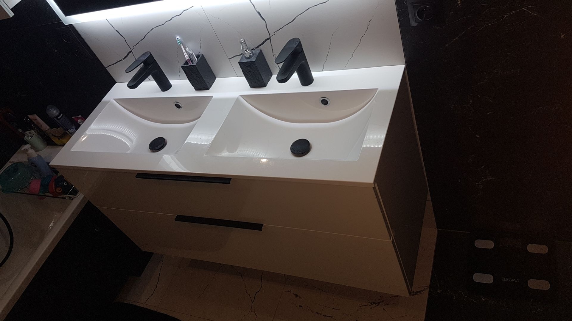 Podwójna, biała umywalka z czarnymi bateriami i czarnymi akcesoriami łazienkowymi na tle marmurowej ściany z oświetleniem LED.