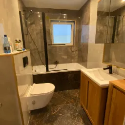 Nowoczesna łazienka z marmurowymi płytkami, drewnianą szafką pod umywalką, wiszącą toaletą i kabiną prysznicową z czarnymi ramami, oświetlona górnym światłem.