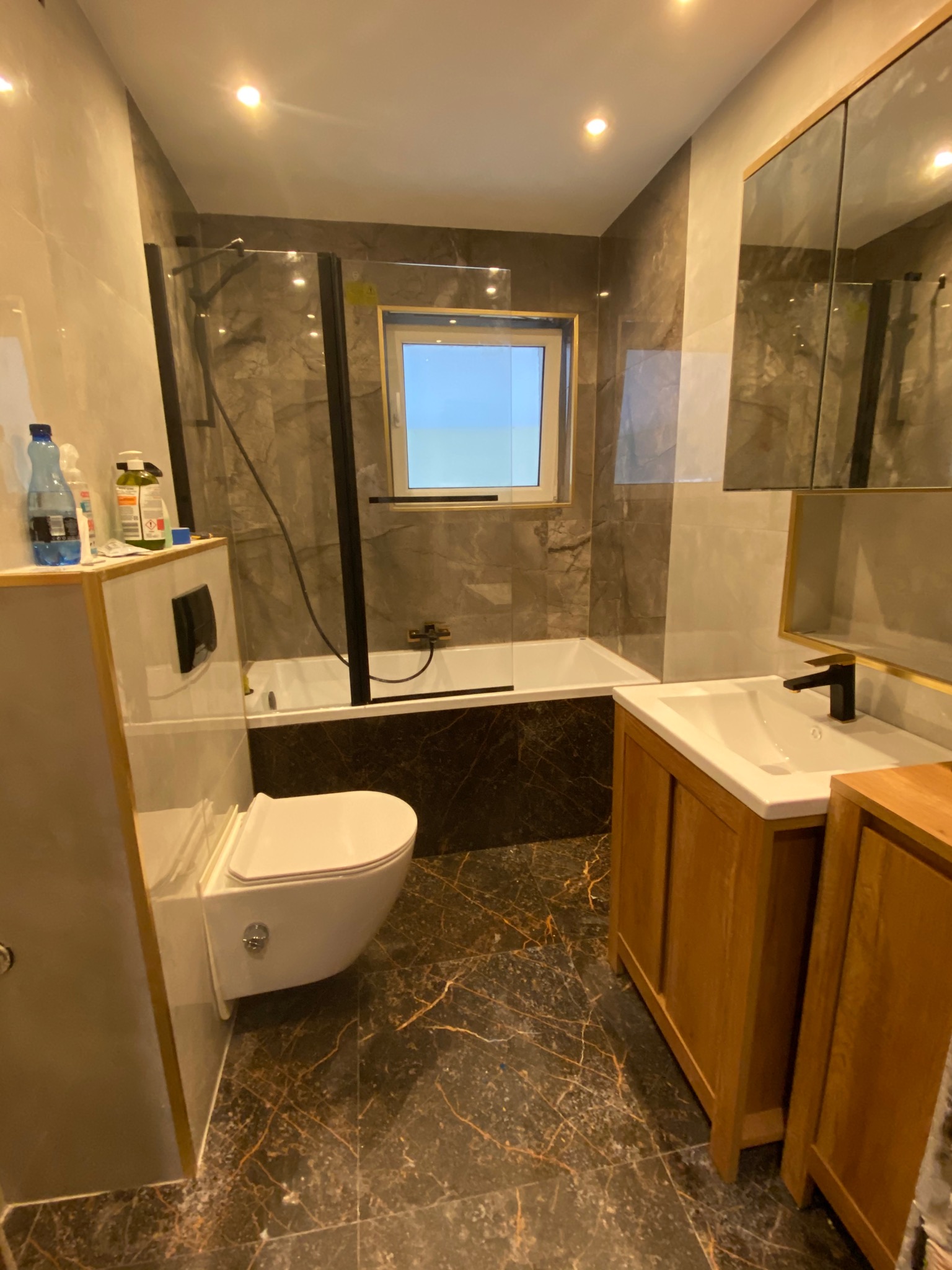 Nowoczesna łazienka z marmurowymi płytkami, drewnianą szafką pod umywalką, wiszącą toaletą i kabiną prysznicową z czarnymi ramami, oświetlona górnym światłem.