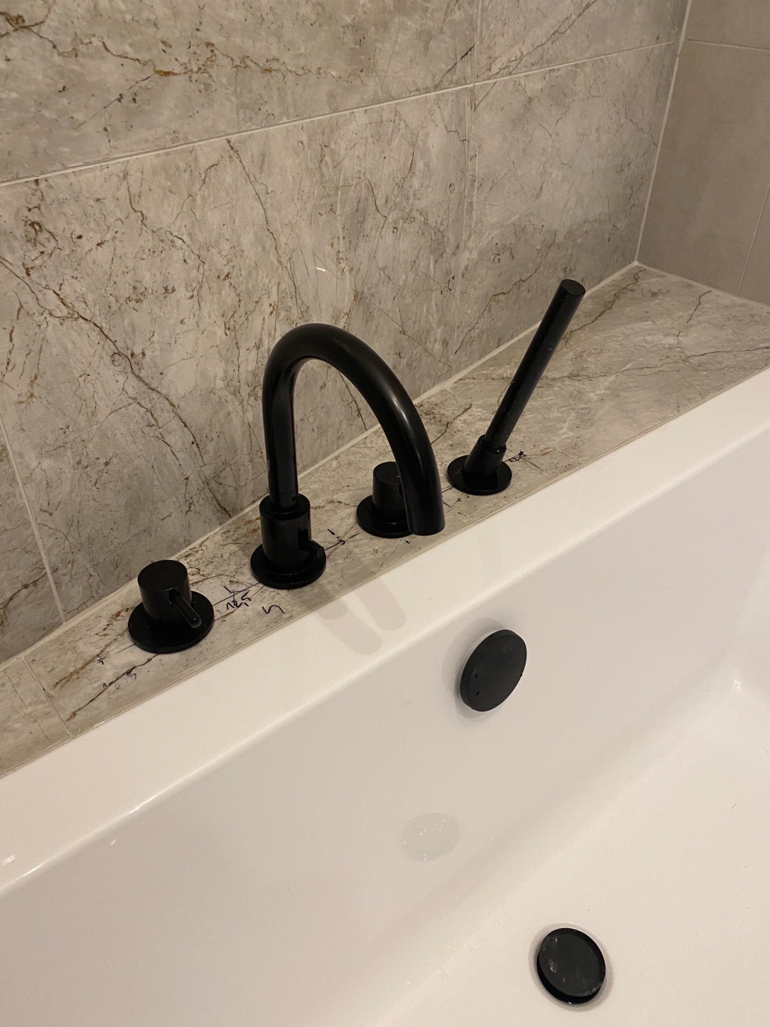 Czarna bateria wannowa zamontowana na marmurowej obudowie wanny, widoczne ślady ołówka po montażu, płytki imitujące marmur na ścianie.